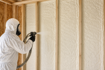 Worker in white protective suit spraying foam insulation into wall studs.