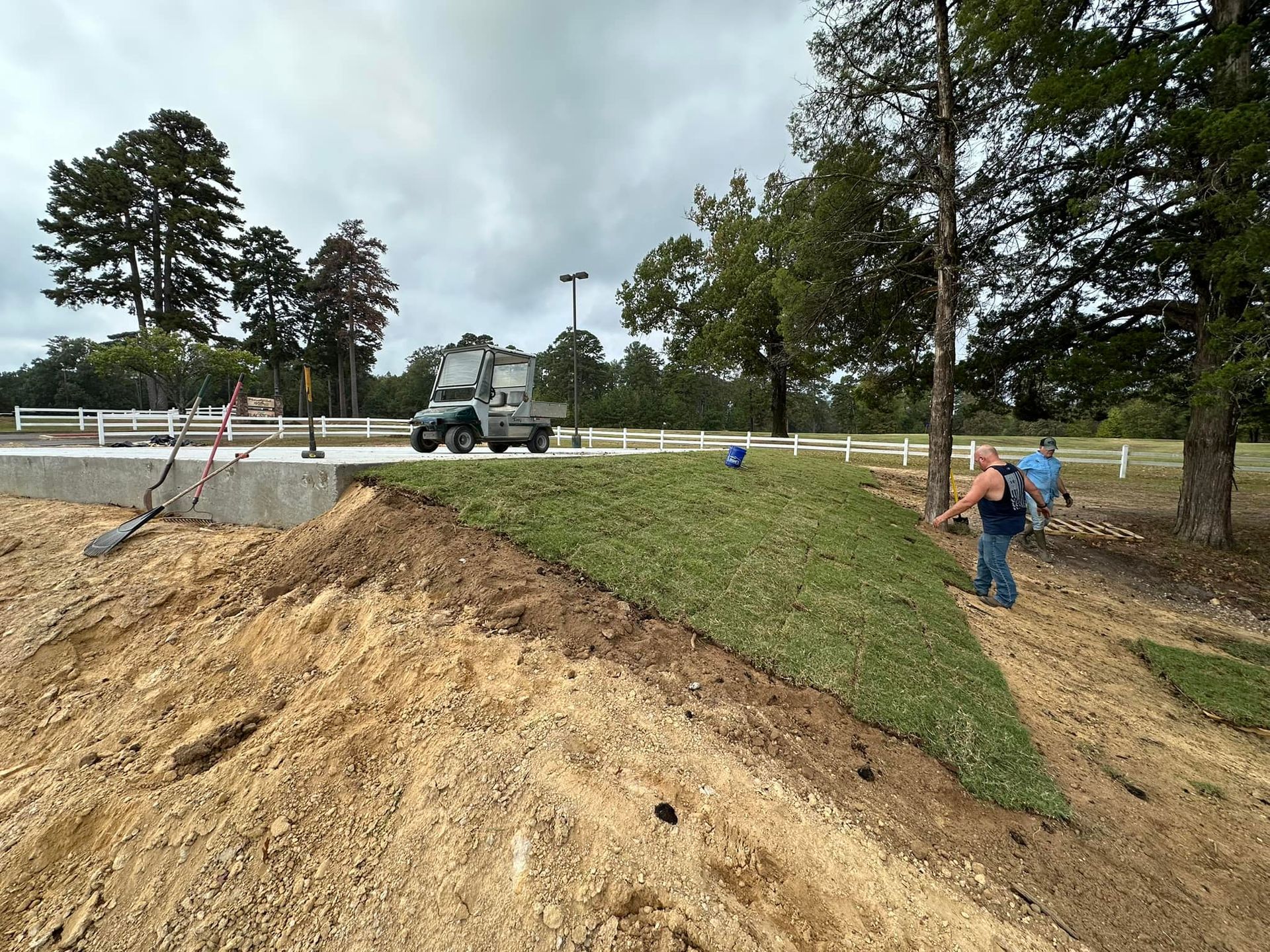 Workers installing sod on a dirt hill. A utility vehicle sits nearby. Overcast sky, outdoor setting.