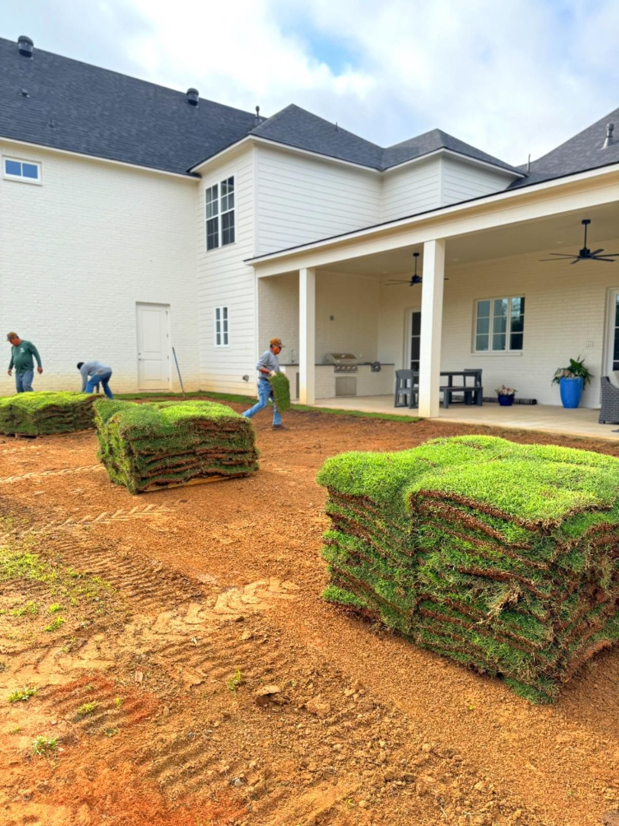 Workers installing sod in a yard near a white house with a covered patio. Green sod stacked and being placed on brown dirt.