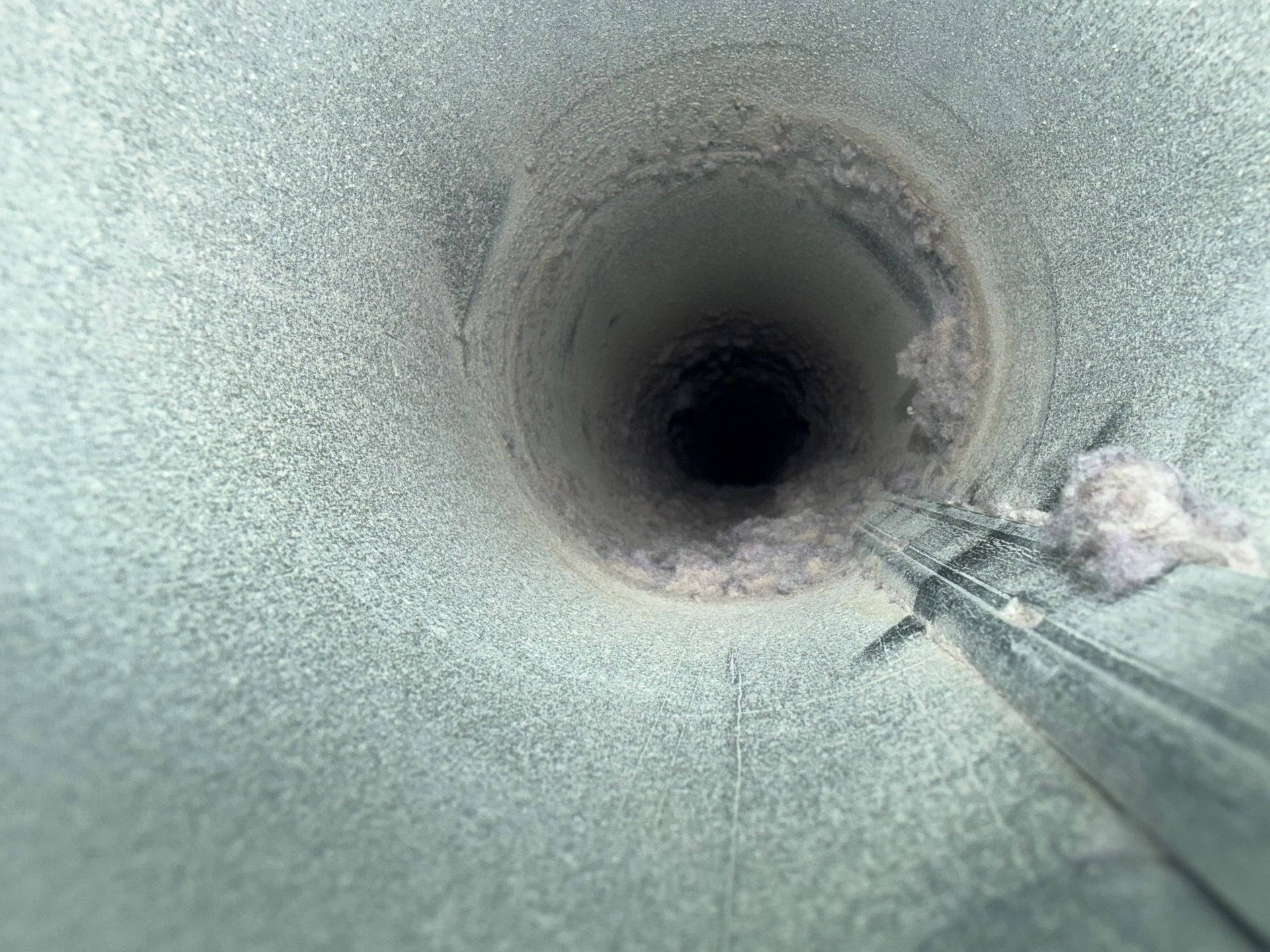 Looking up inside a narrow pipe toward a dark opening, with a cleaning tool at the edge