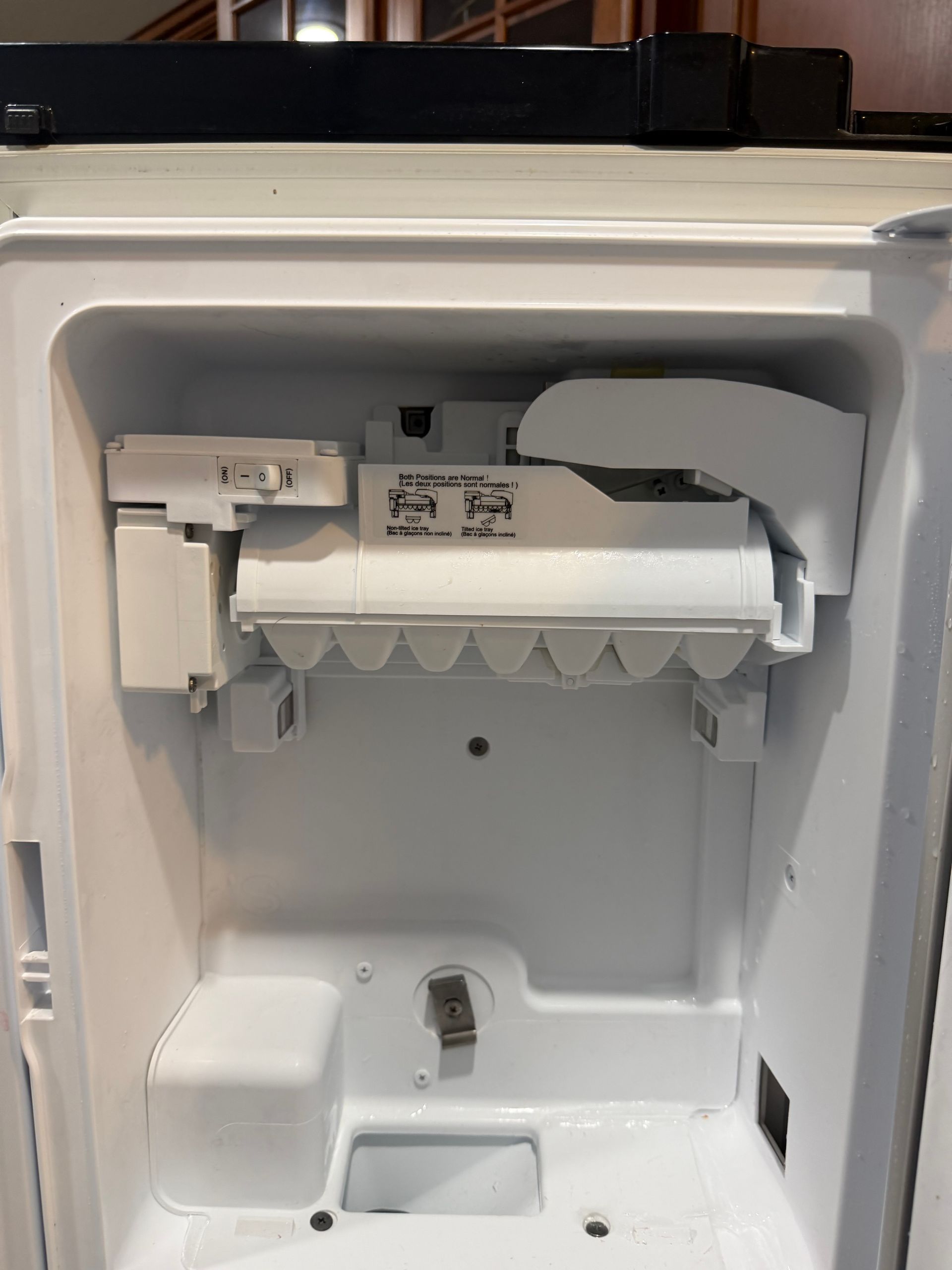 Open refrigerator ice maker compartment with white plastic components and bin cavity.