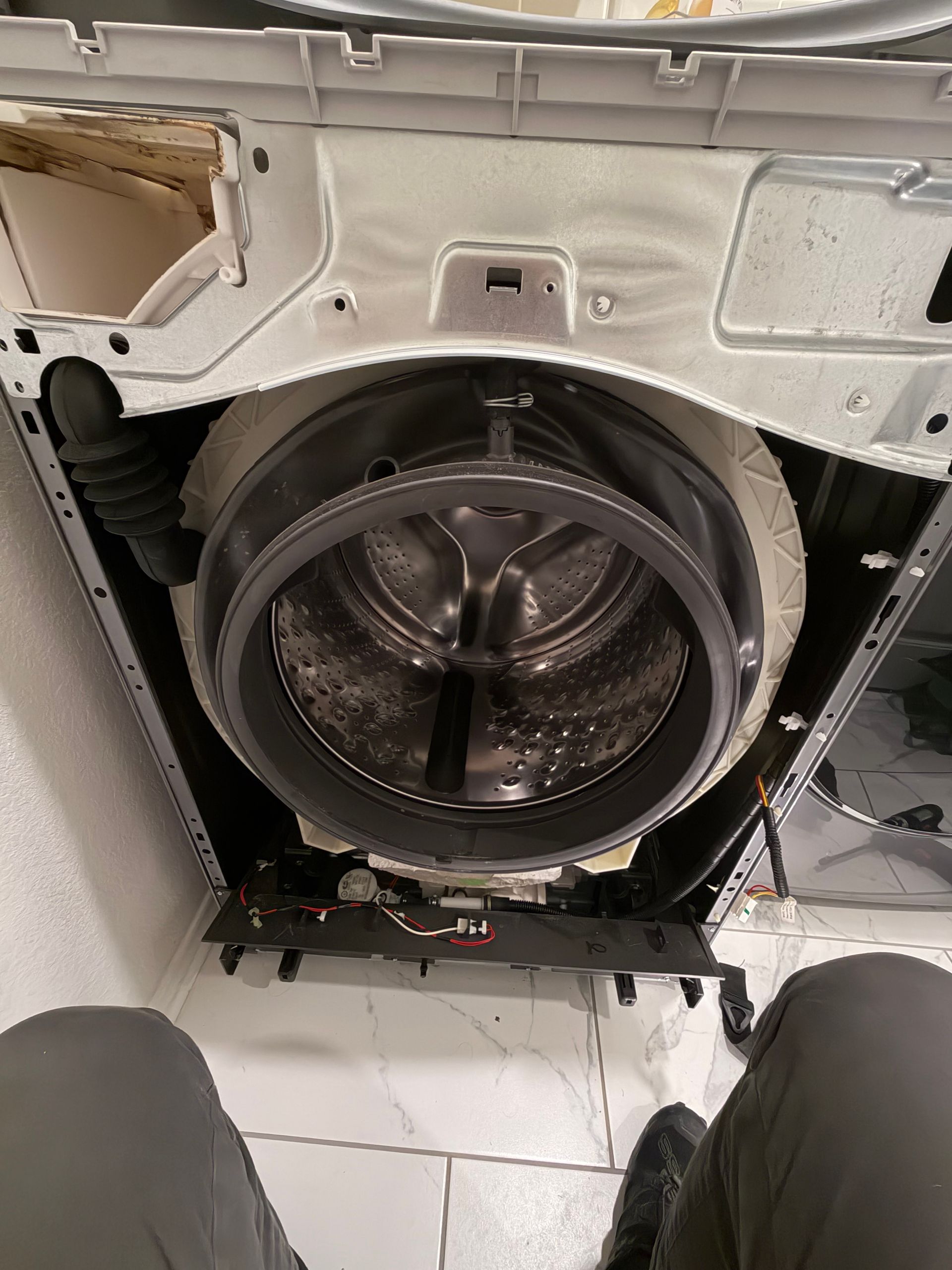 Open washing machine with drum exposed during repair, showing internal metal parts and white exterior panel.