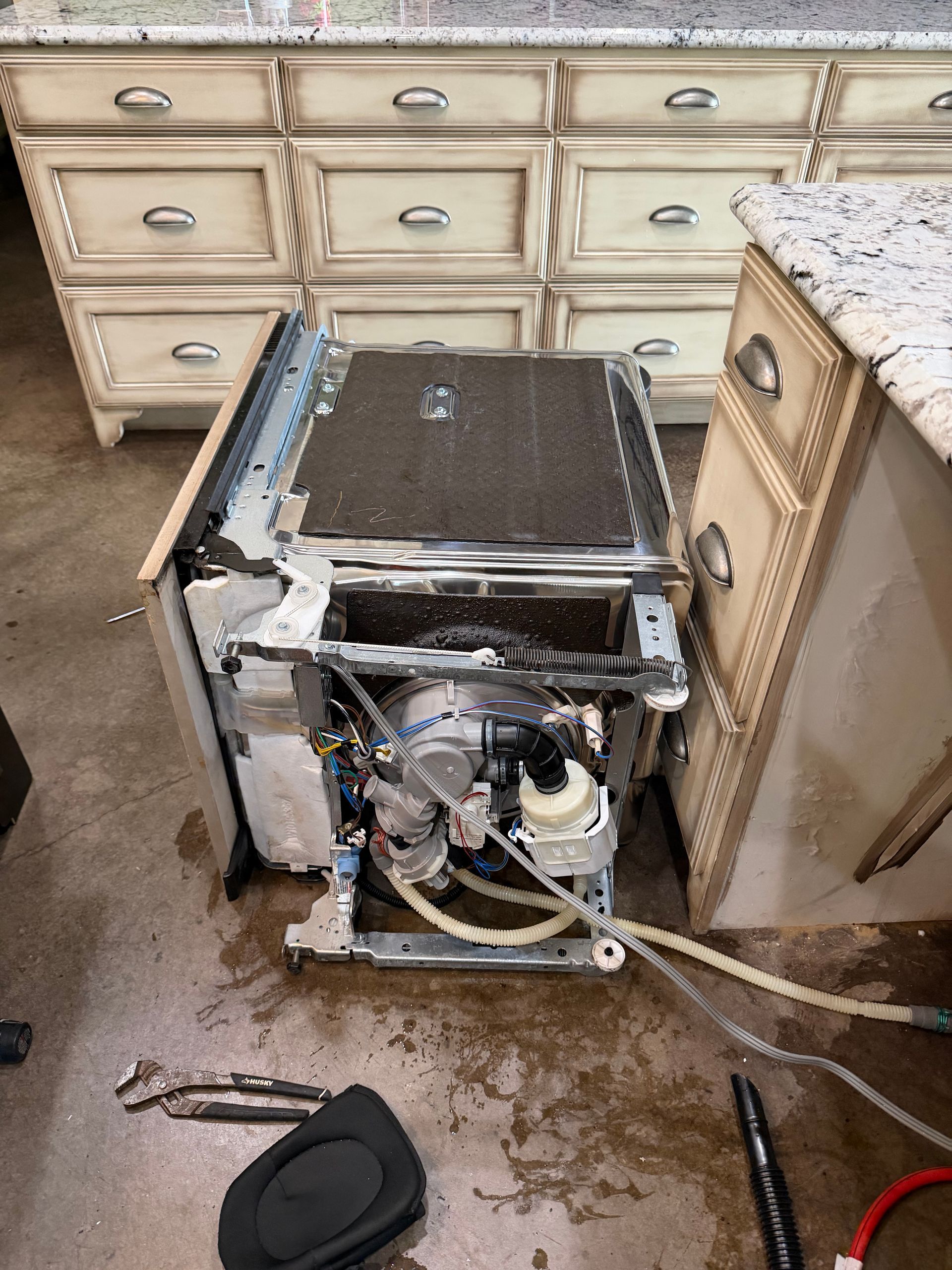 Damaged dishwasher in a kitchen, leaking water with its front panel removed.