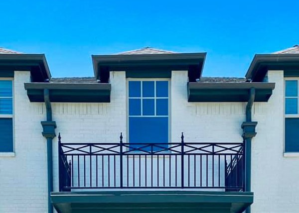 White brick building with a black balcony, blue window, and green trim against a clear, blue sky.