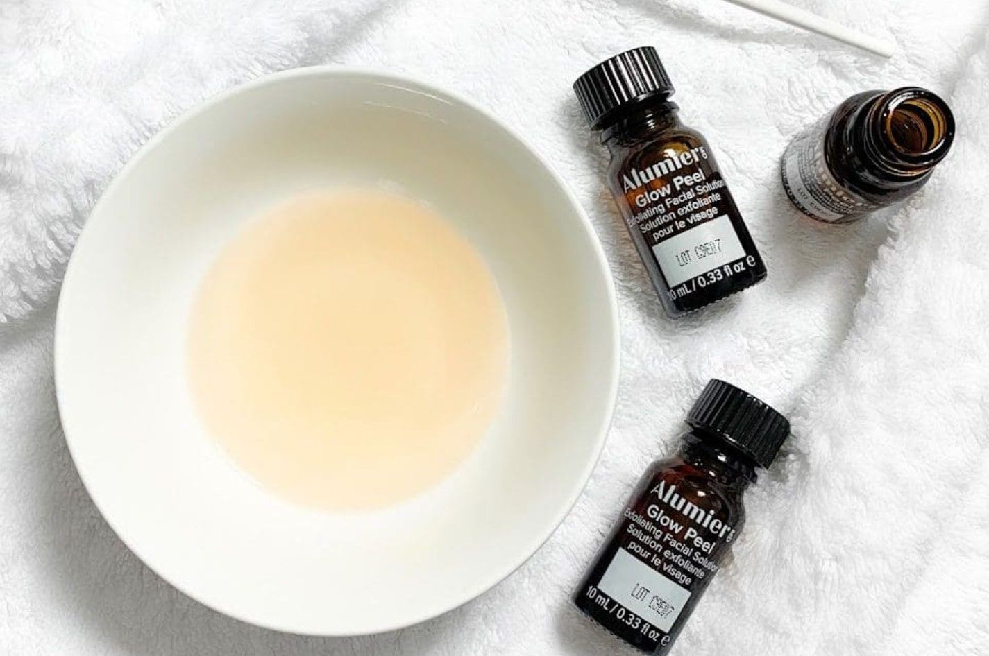 White bowl of pale liquid with three small black essential oil bottles on a white towel