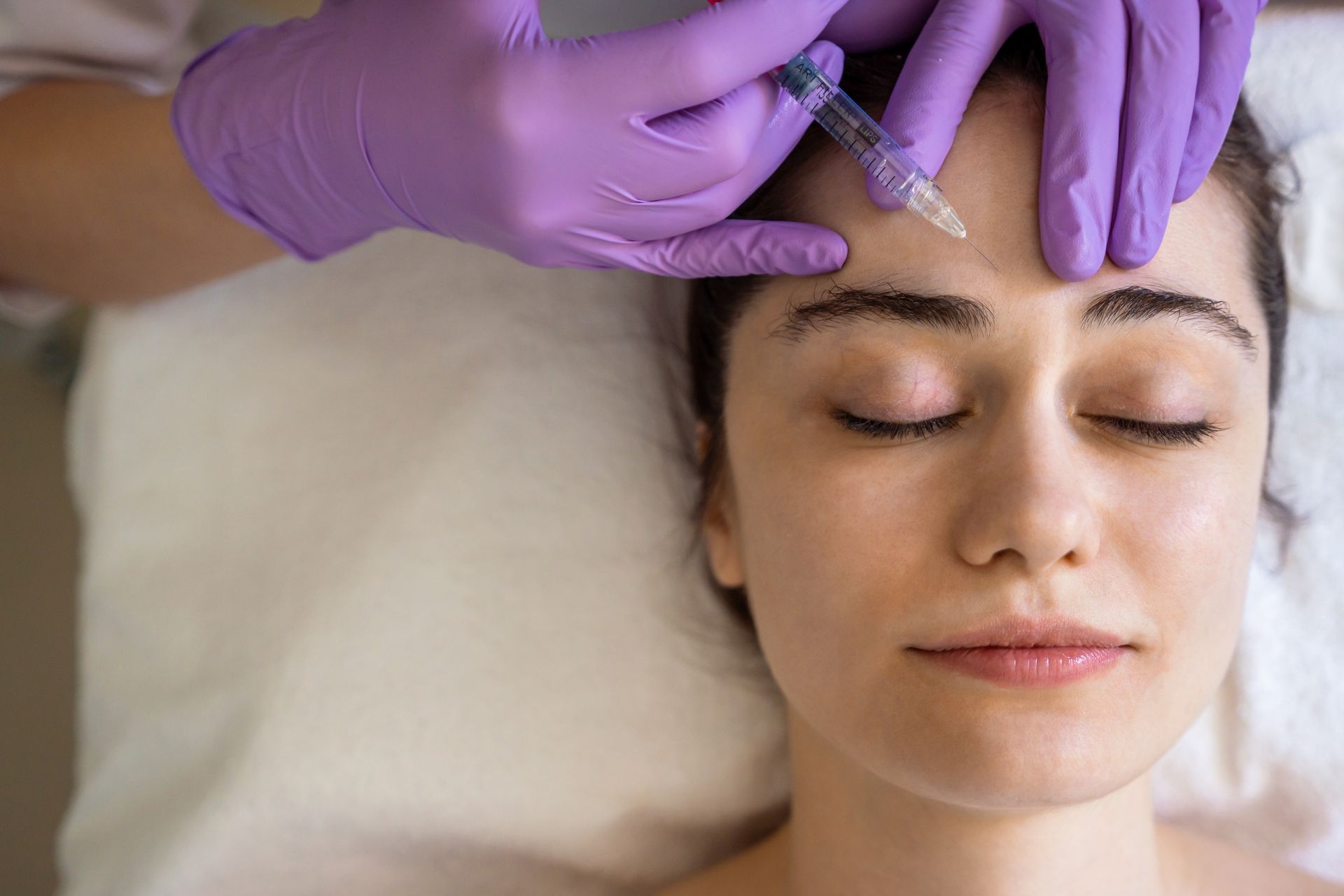Gloved hands injecting a woman’s forehead as she lies with eyes closed on a treatment bed
