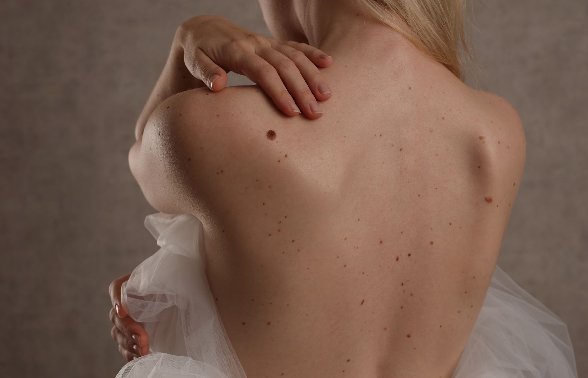 Woman with bare back and hand on shoulder, draped in white fabric against a neutral background