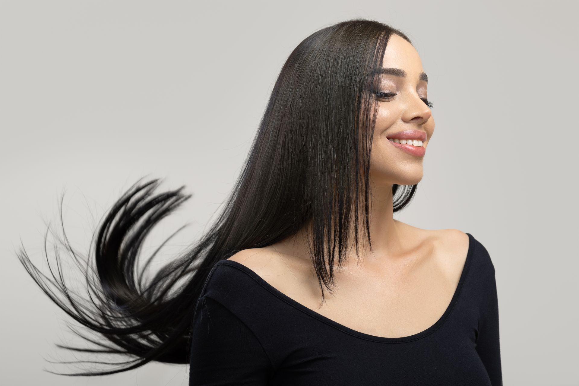 Smiling woman with long black hair in a black top, hair flowing against a light gray background