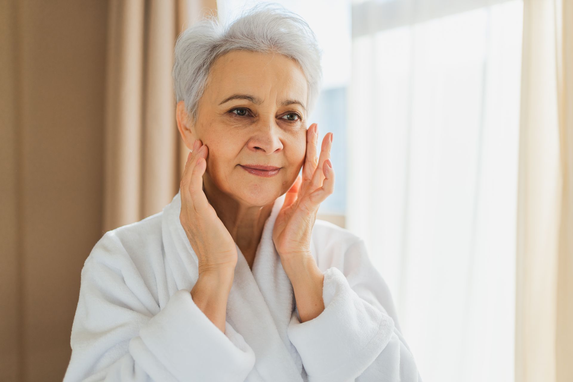 Person in a white robe gently touching their face in a softly lit room