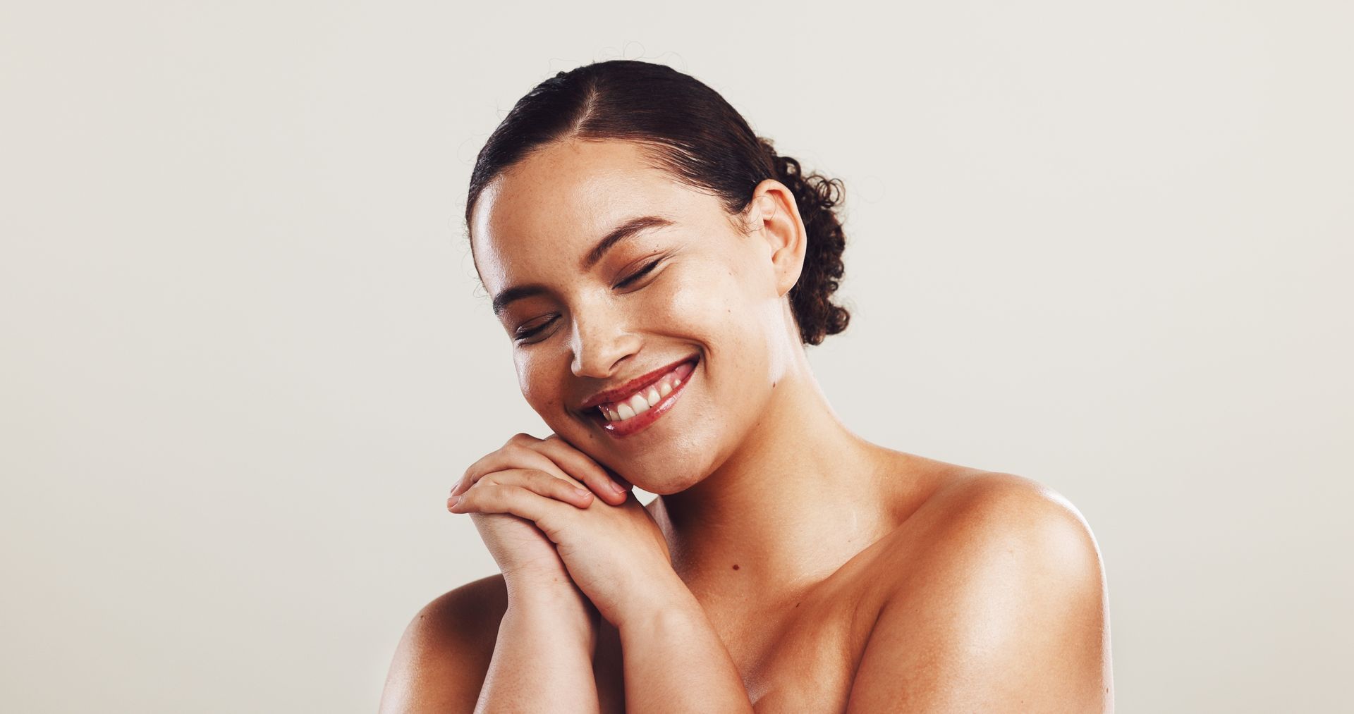 Smiling woman with closed eyes resting her head on clasped hands against a light background