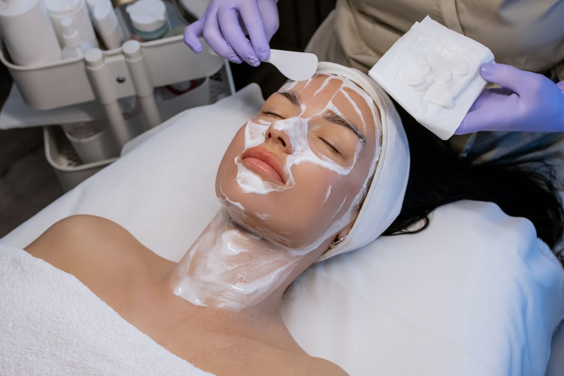 Person applies a facial mask with a brush while another lies on a treatment bed