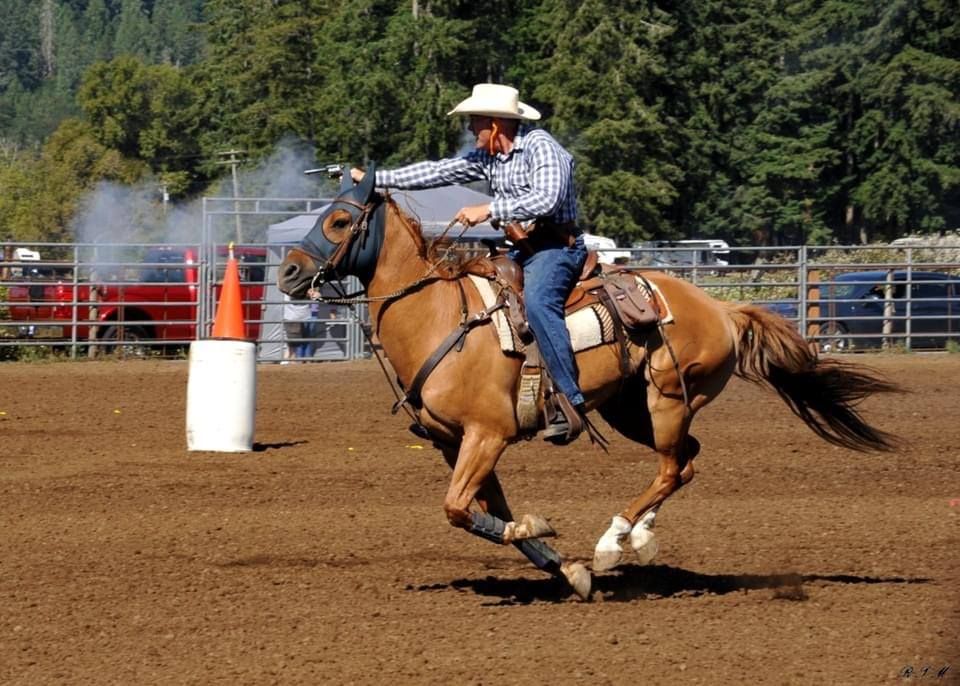 A rider on a brown horse at full gallop in a dirt arena, passing a white barrel during a mounted shooting event.