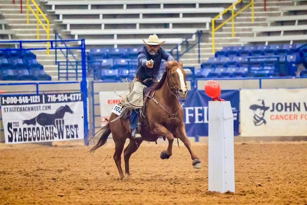 A person on horseback aiming a firearm at a red balloon atop a white pedestal in an indoor arena.