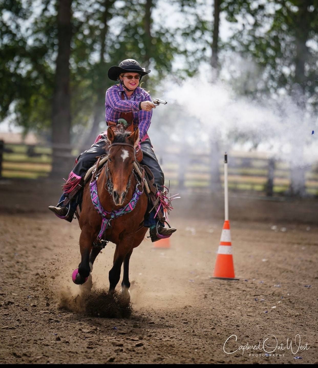 A person in a cowboy hat rides a brown horse, firing a blank pistol at a target in a dirt arena.