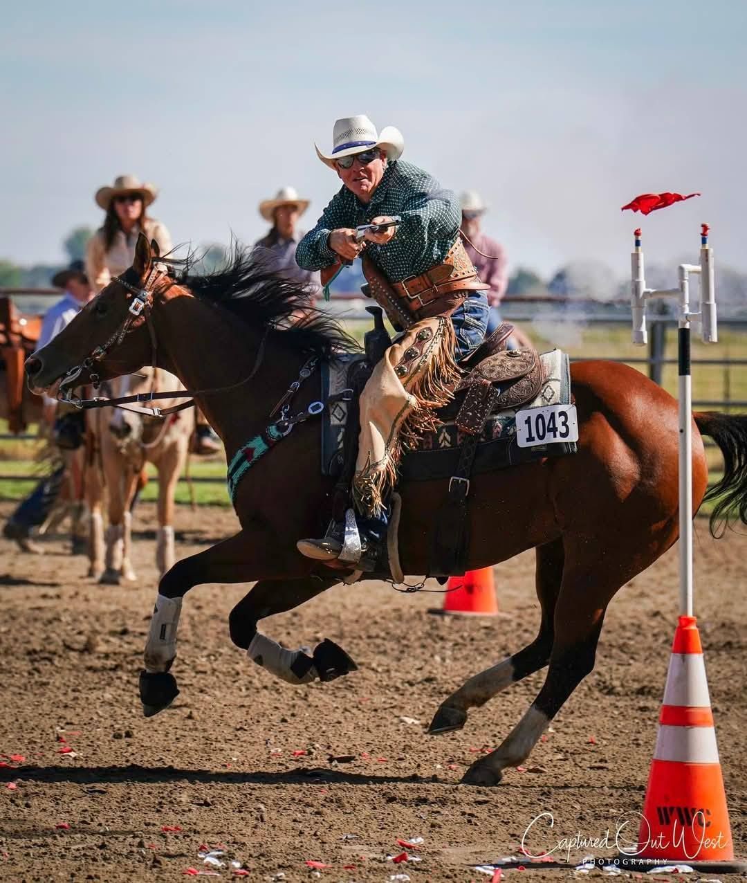 A horseback rider in Western gear aiming a pistol at a target in a sandy arena during a mounted shooting competition.