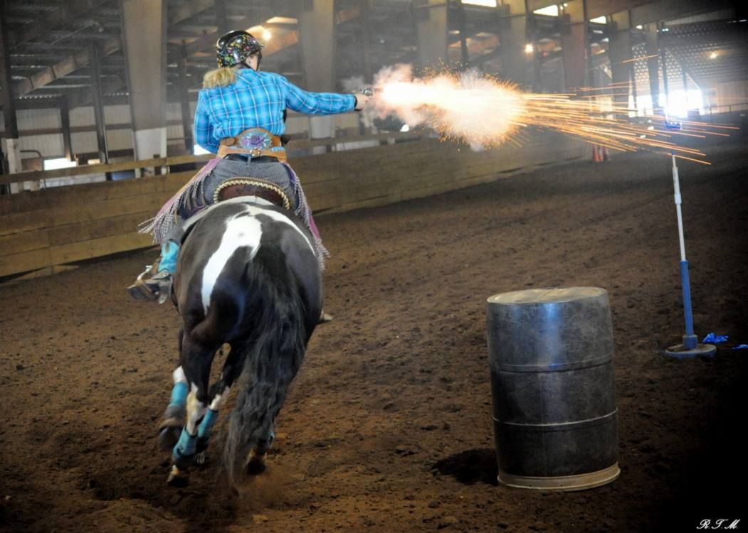 A mounted rider in a blue plaid shirt fires a gun at a target while riding past a barrel in an indoor arena.