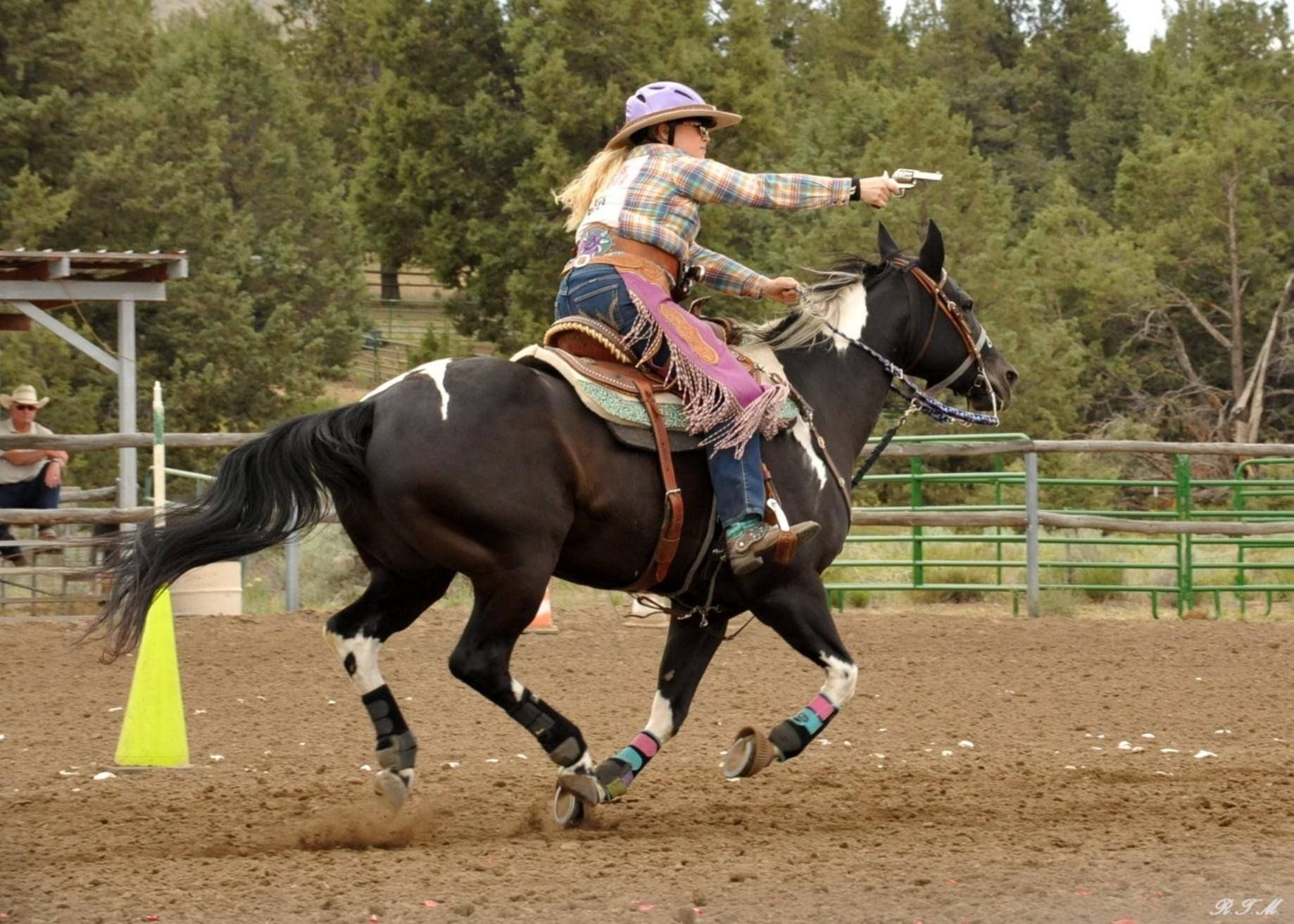 A person in Western attire rides a black and white horse at a gallop, pointing a handgun toward a target in an arena.