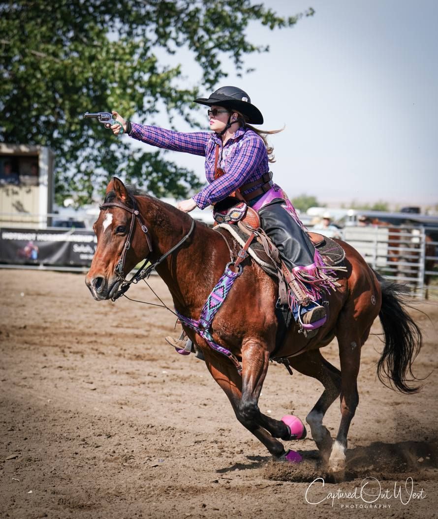 A rider in a purple plaid shirt and black hat aiming a pistol while riding a brown horse in an arena.