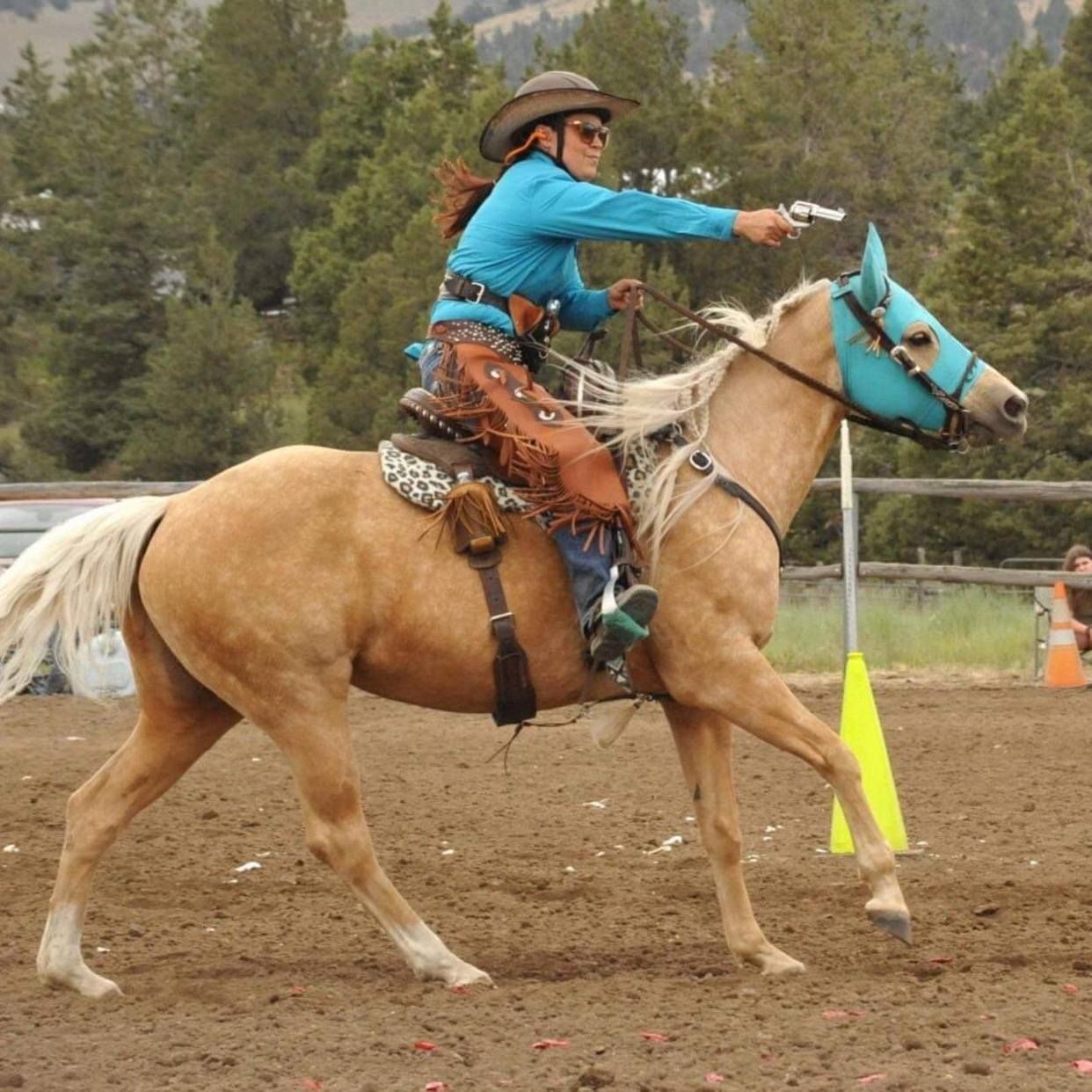A rider in a blue shirt and hat aims a pistol while mounted on a palomino horse with a blue fly mask in a riding arena.