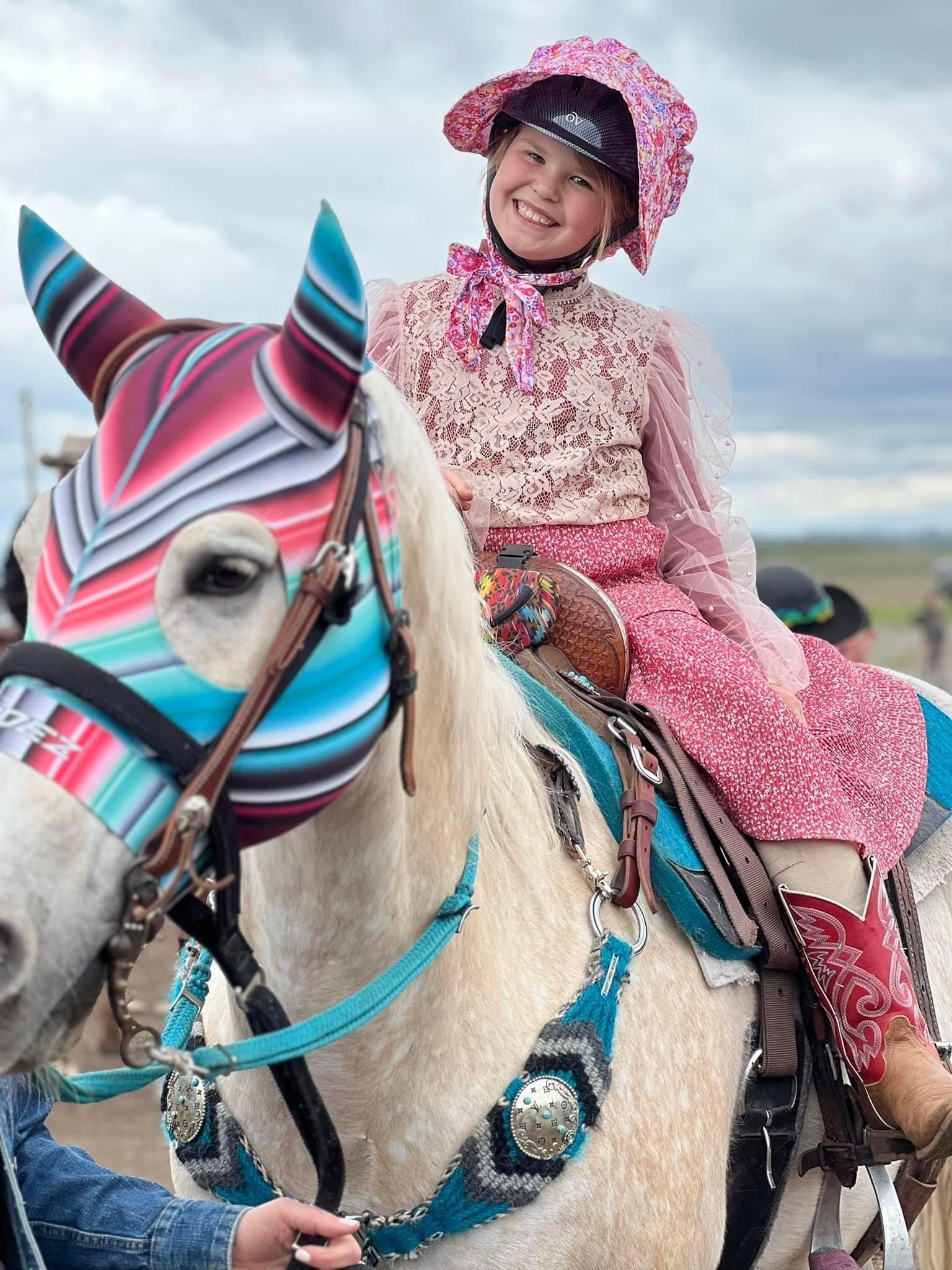 A rider in a pink bonnet and lace top sits on a white horse wearing a colorful, striped mask and turquoise tack.