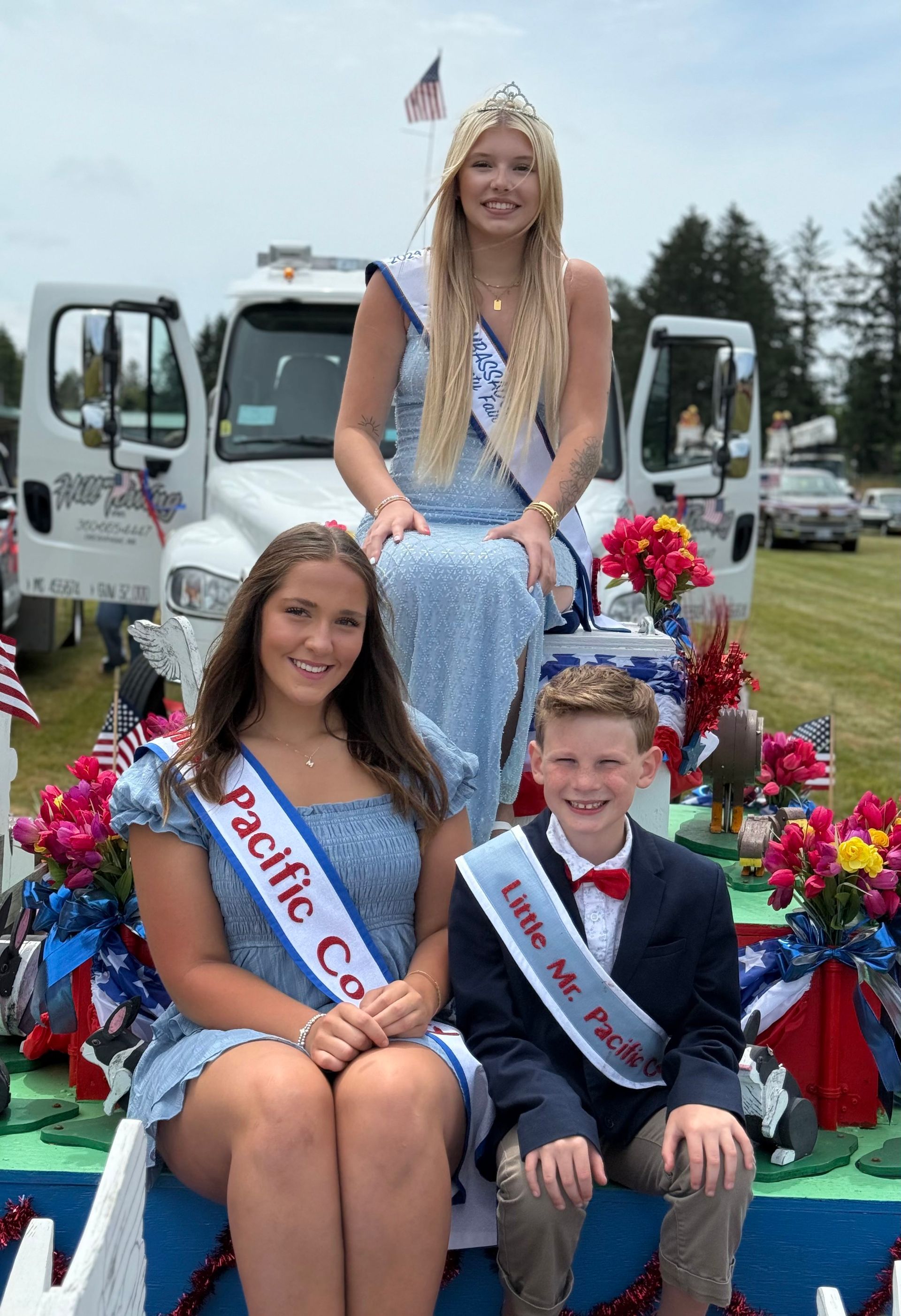 Three people in sashes sit on a parade float decorated with flowers and American flags.