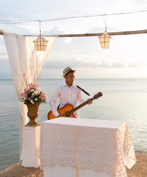 Man plays guitar at an outdoor seaside wedding ceremony; white tablecloth, floral arrangement, and hanging lanterns.