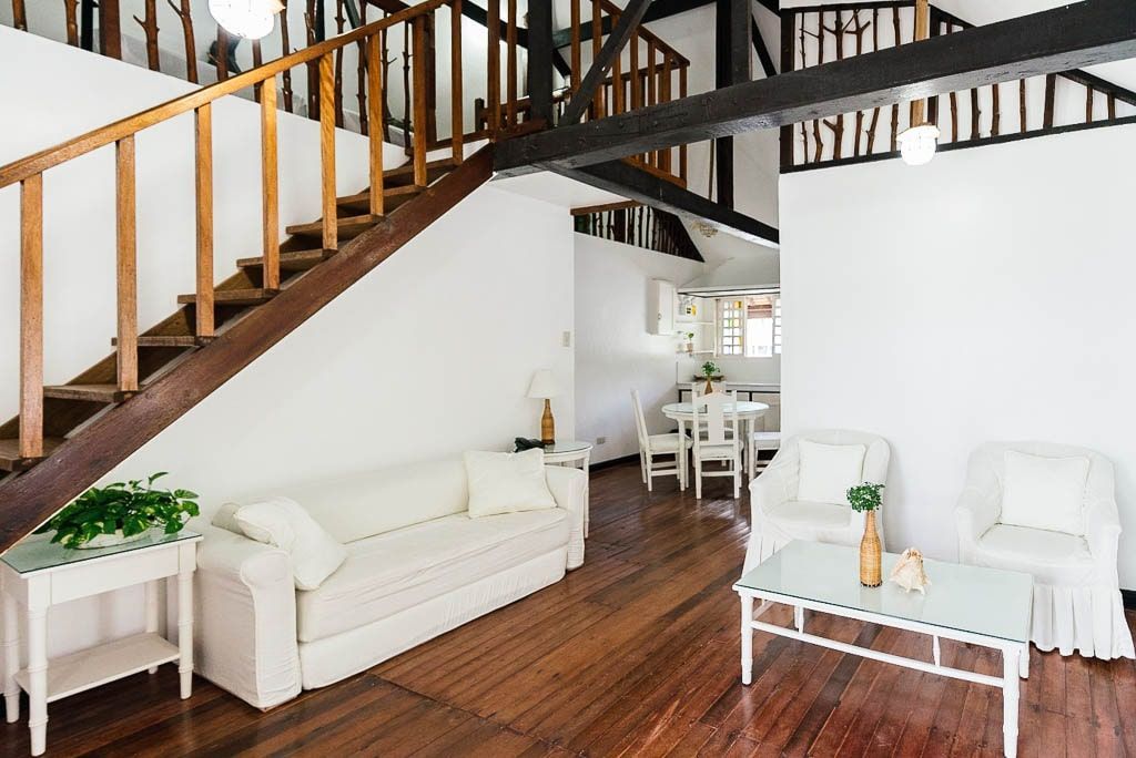 Living room with stairs, white sofa and chairs, wooden floors and beams.
