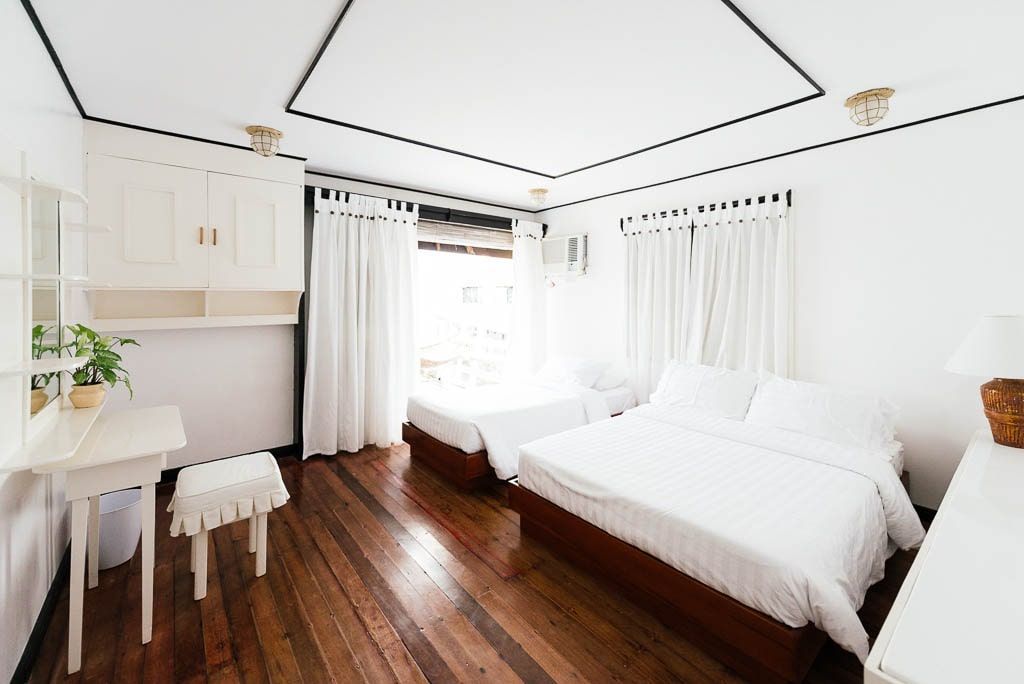 Bright bedroom with two beds, white linens, hardwood floors, white curtains, and a desk.