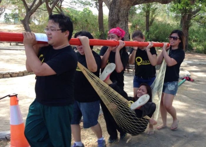 Six people carry a person in a net suspended from a pole outdoors.