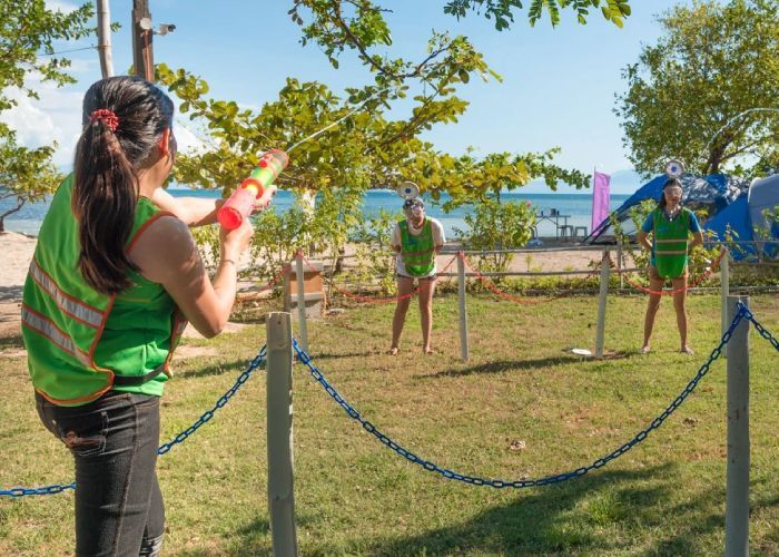 Woman aims water gun at two people wearing green vests, playing on a lawn near a beach.
