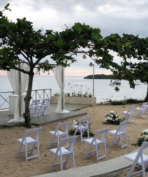 Wedding ceremony setup on a beach: white chairs, draped altar, ocean view, cloudy sky.