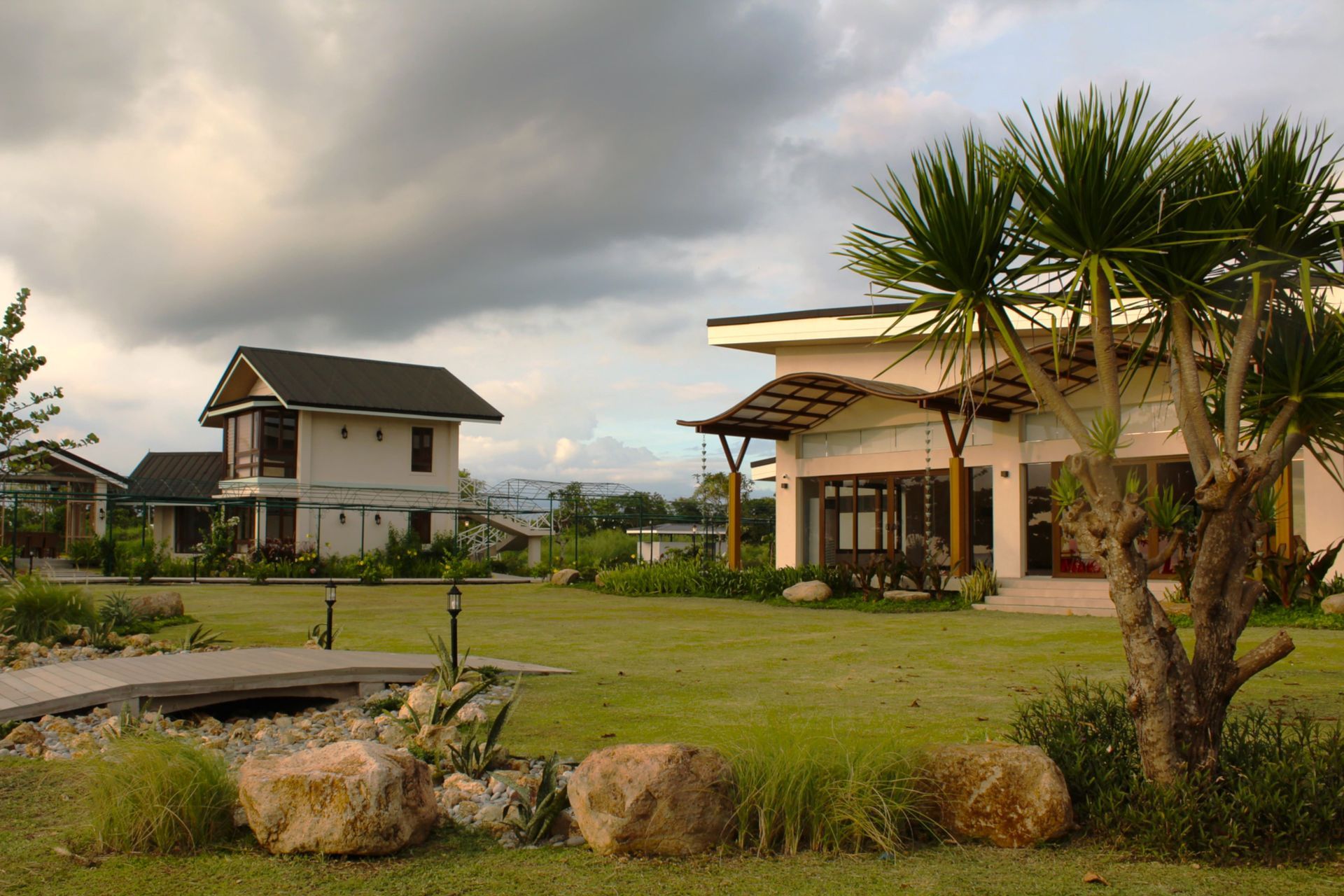 Lush green lawn with a bridge, large rocks, and several houses under a cloudy sky.