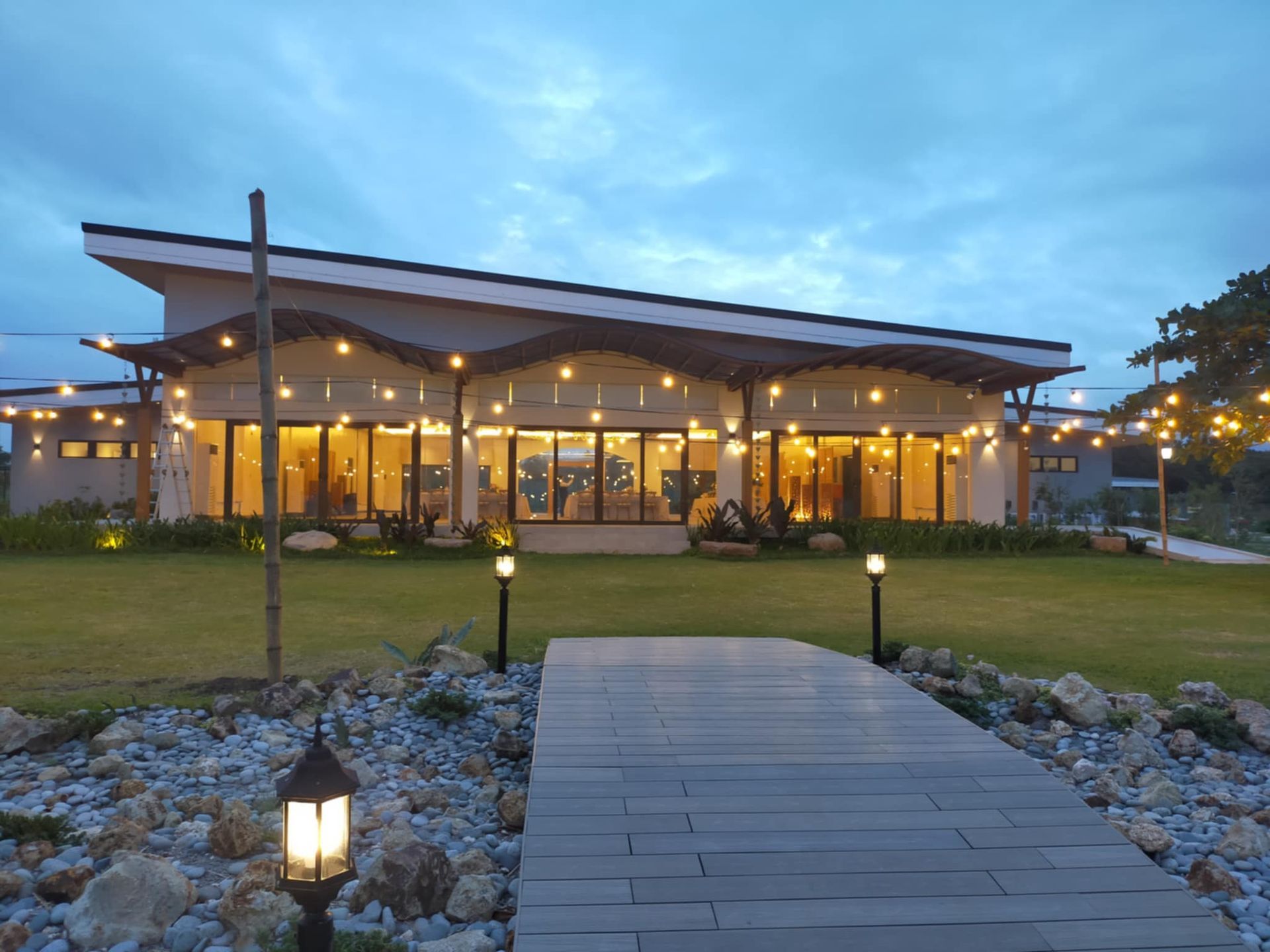 A building with string lights at dusk. A wooden pathway leads to the glass-fronted structure.