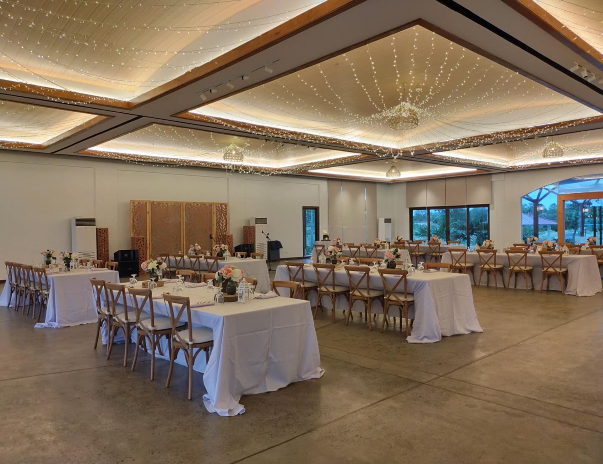 Empty event hall with tables, chairs, and stage. Neutral colors, overhead lighting, and white tablecloths.