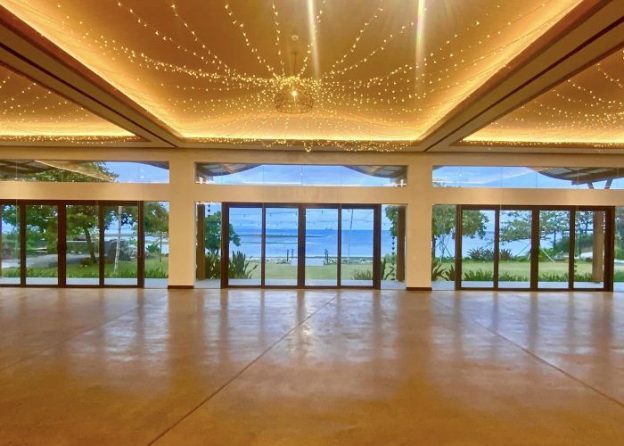Empty room with glass doors facing the ocean, wood floor and ceiling with string lights.