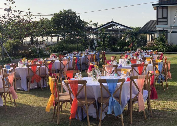 Outdoor reception tables with colorful fabric on chairs and centerpieces in a grassy area.