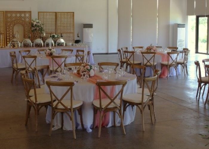Banquet hall with round tables set for a meal. Tables have pink accents and surrounded by cross-back chairs.