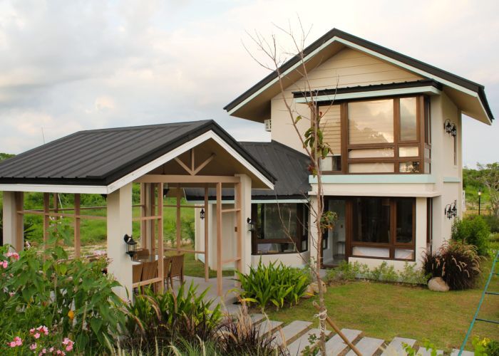 Two-story house with black roof, large windows, and an adjacent covered structure; surrounded by greenery.