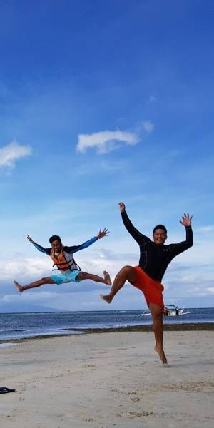 Two people jumping on a sandy beach under a blue sky. One person wearing blue/orange, the other wearing red/black.