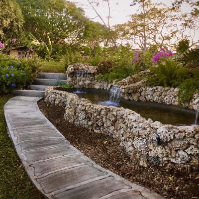 Stone pathway leads past tiered water feature with cascading waterfalls, lush garden setting.