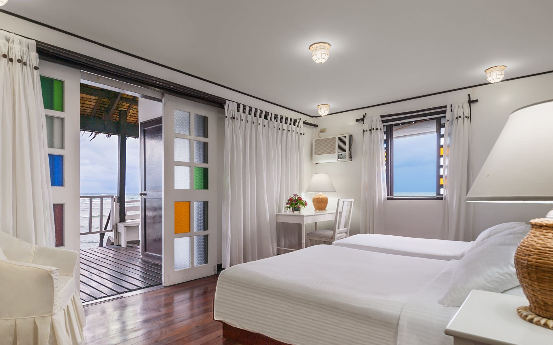 Bright bedroom with two beds, balcony access, white decor, and colorful stained glass accents.
