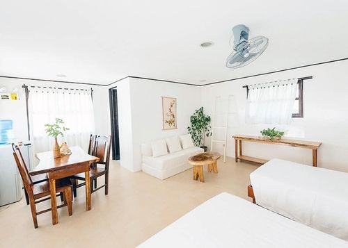 Bright, white open-concept living space with dining table, sofa, two beds, and decorative plants.