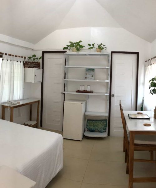 A bright white room with a bed, desk, shelving, refrigerator, and table with chairs.