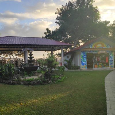 Park scene with fountain, covered area, and small building with colorful artwork; green grass and cloudy sky.