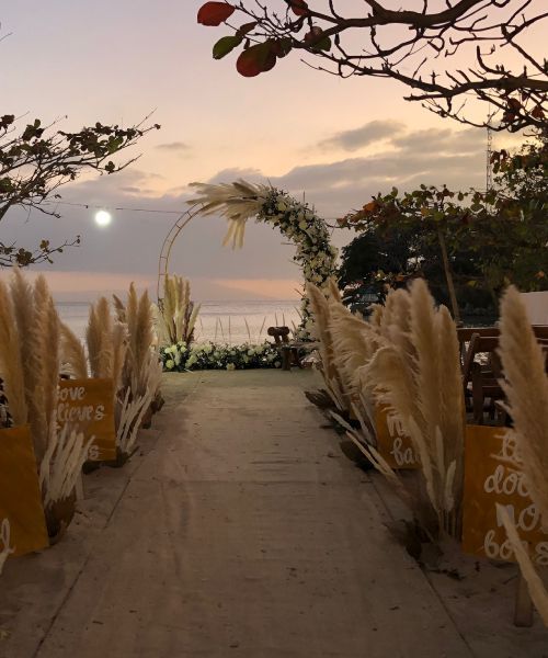 Beach wedding setup with pampas grass, arch, and ocean view at sunset.