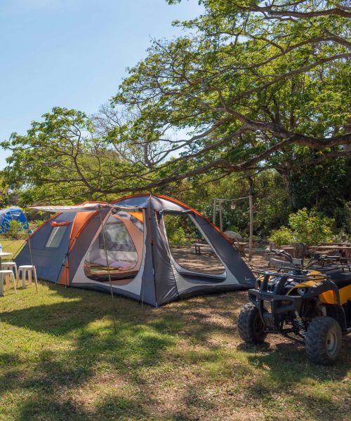 Large camping tent set up on a grassy area, under a tree. A yellow ATV is parked nearby.