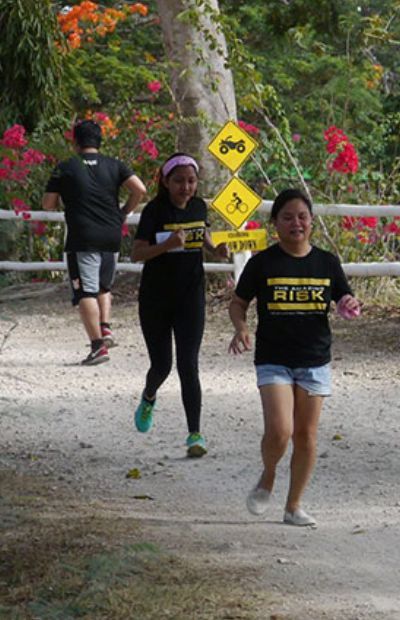 Three people running on a dirt path, near a sign, trees, and flowers.