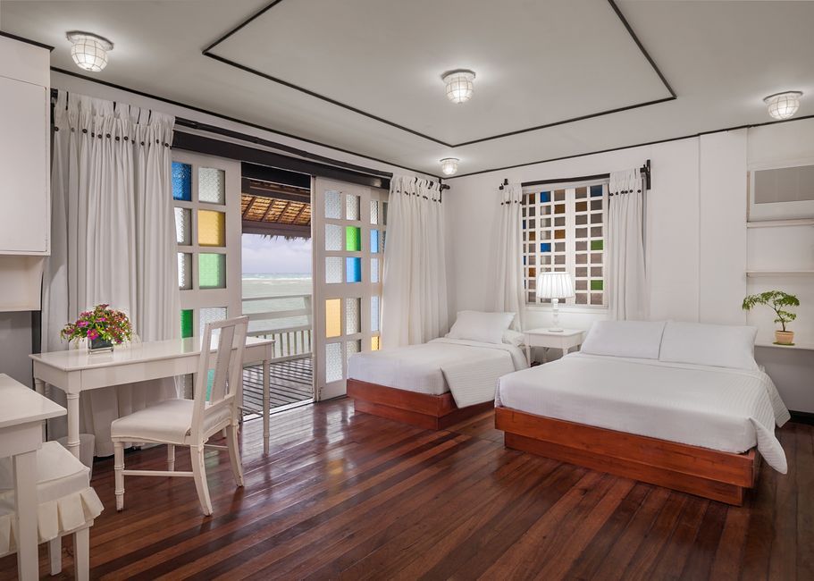 Bright hotel room with wooden floors, two beds, a desk, and a balcony with colorful window panes.