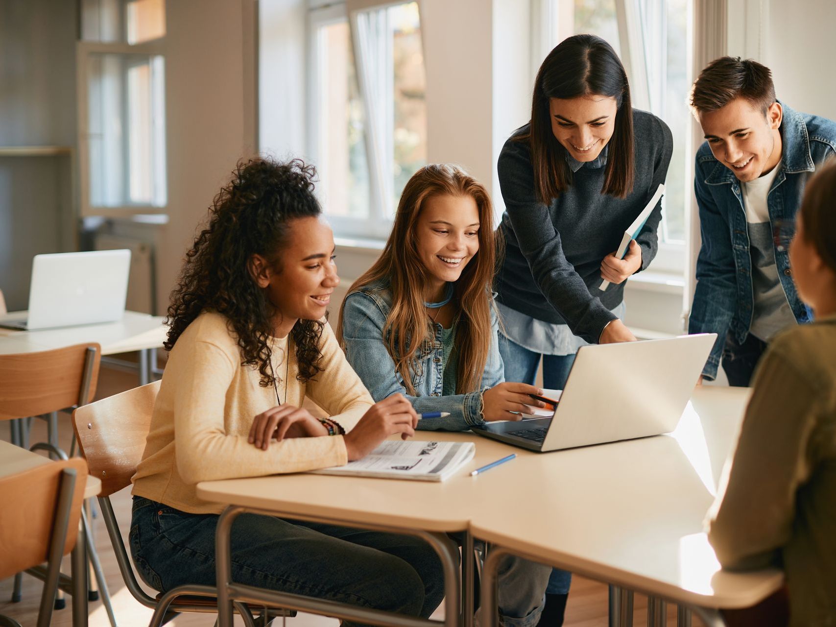 Students around a laptop, smiling, in a classroom setting.