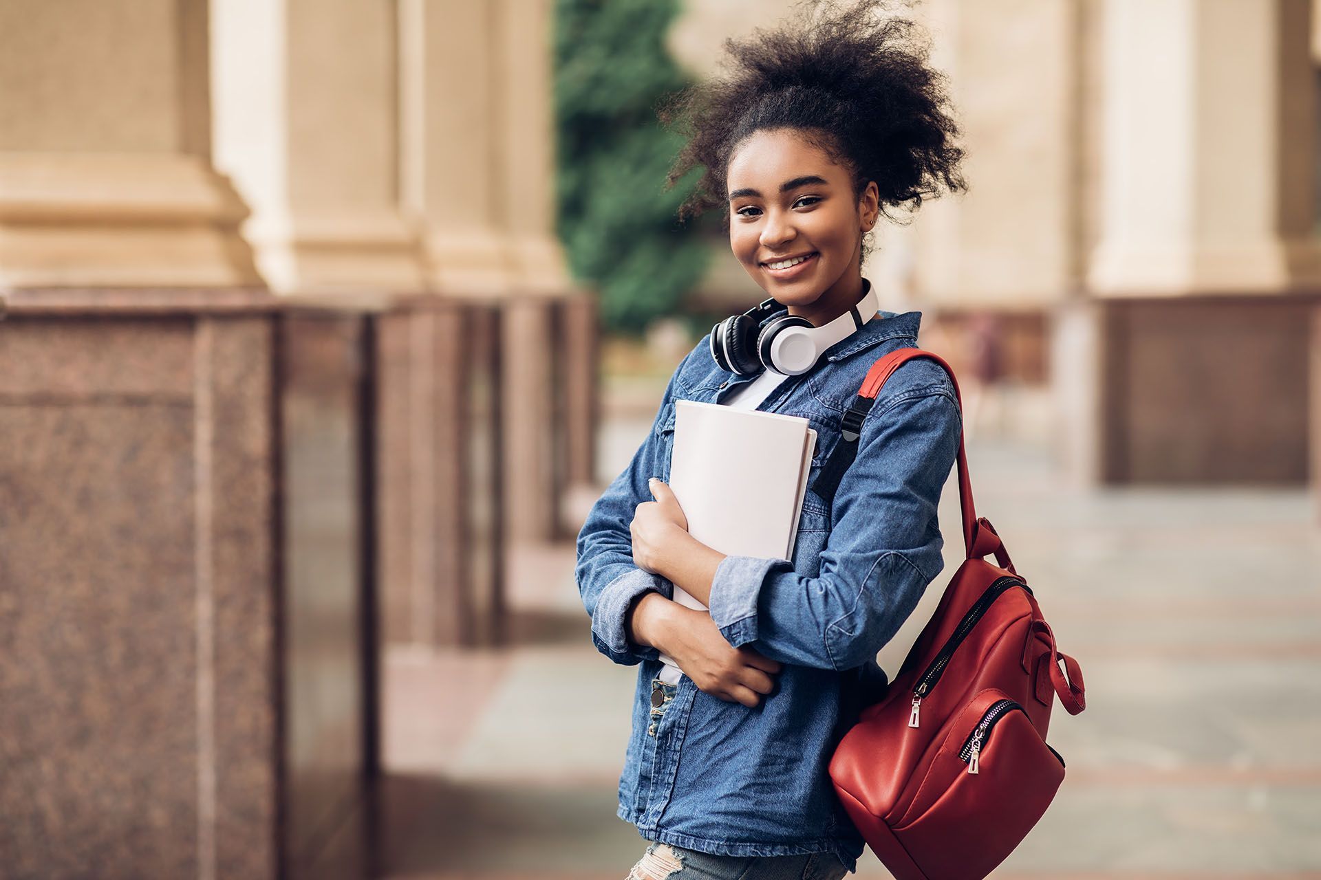 Smiling person with curly hair, headphones, red backpack, holding books, near a building.